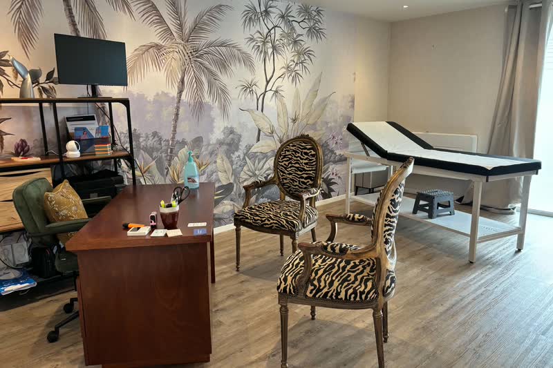 Salle de consultation : bureau, deux fauteuils et table d'examen devant un papier peint de palmiers